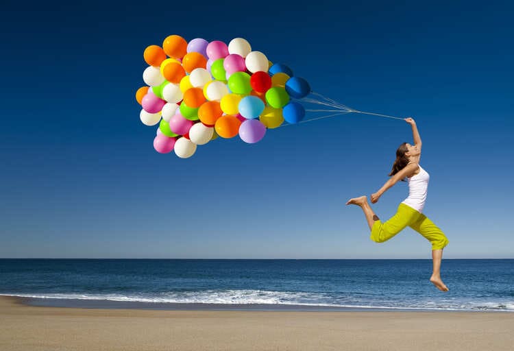 Frau mit vielen bunten Luftbalon an der Schnur, läuft am Strand und hüpft fröhlich in die höhe