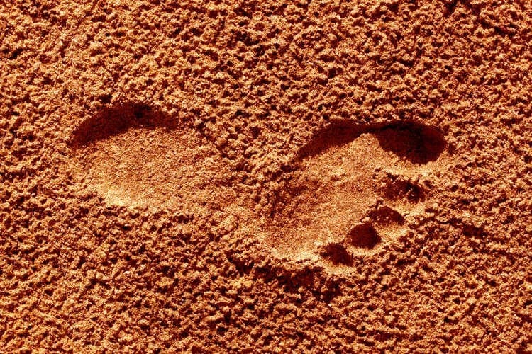 Zwei barfuß hinterlassene Fußabdrücke im Sand, die eine tiefe Verbindung zur Erde und das bewusste Erleben des Moments symbolisieren. Das Bild vermittelt das achtsame Gehen und das Fühlen des Bodens unter den Füßen. Im hier und jetzt leben