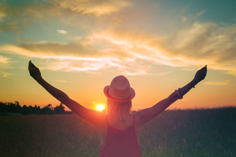 Rückansicht einer Frau mit ausgebreiteten Armen, die bei Sonnenuntergang in einem Feld steht und den Himmel betrachtet. Das Bild vermittelt Offenheit, Freiheit und die Entfaltung des eigenen Selbstwerts. Erkenne deinen Wert