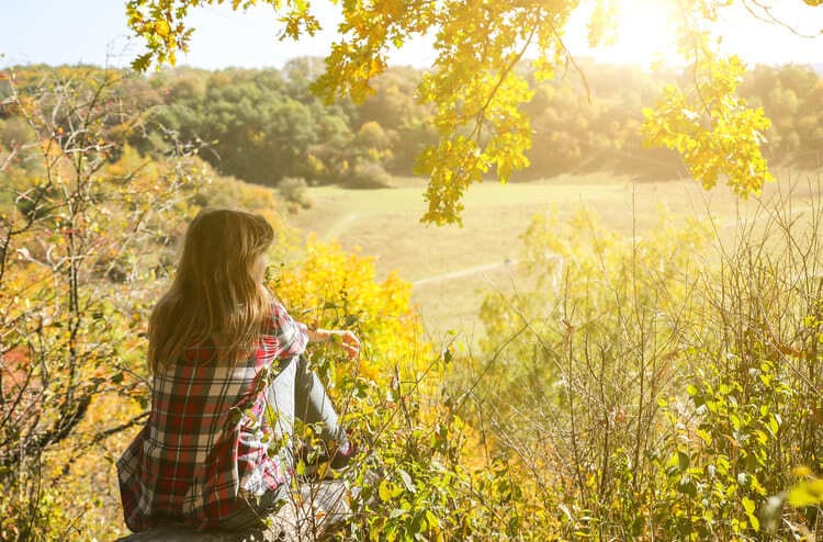 Frau sitzt inmitten herbstlicher Natur und blickt nachdenklich in die weite Landschaft, umgeben von warmem Sonnenlicht und grünen Blättern – Symbol für Ruhe, Reflexion und innere Akzeptanz. Rdaikale Akzeptanz